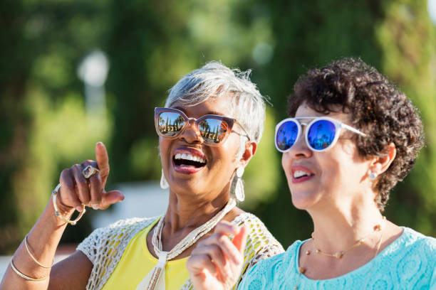 A beautiful senior African-American woman in her 70s and her Hispanic friends, standing outdoors on a sunny day, wearing sunglasses, laughing. The focus is on the older woman.