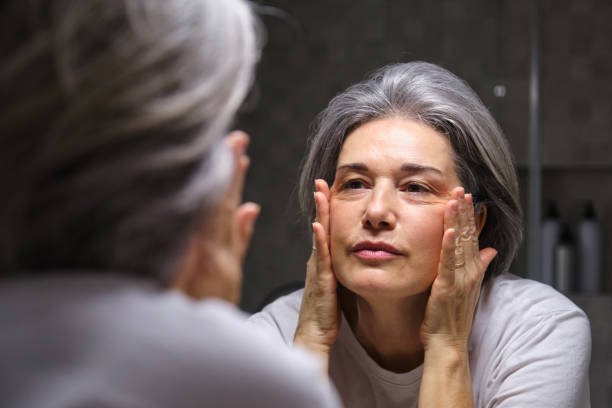 Mature woman with gray hair looking at her wrinkles in the mirror, worried about aging